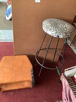 Both stools together, showing the tall black metal stool with floral cushioned seat and the brown leather folding stool.