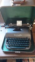 Remington Office Model portable typewriter inside open green felt-lined case with brown leather handle, placed on wooden surface.