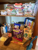 Wide view of lower shelf with stacks of folded dish towels and pot holders, along with cleaning supplies including Dr Teals Epsom salt, Scoop Away cat litter, cleaning liquids, baby wipes, and trash bags.