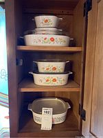 Six Corningware casserole dishes displayed on wooden shelves showing two different floral patterns, clear plastic lids on each.