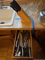 Wooden knife block on kitchen counter, containing several knives with black handles; drawer below holds more knives and utensils.