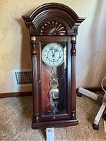 Full view of the wooden pendulum case clock featuring an arched top with carved shell design, decorative columns, and glass door showing pendulum.