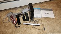 Wide angled photo of black Nintendo Wii console standing vertically with included base stand, motion remote with jacket, nunchuk controller, sensor bar, cables, and manuals on carpeted floor.