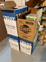 Four large boxes, two Today brand fully closed paper towel boxes, one fully opened Fiora bath tissue box with visible packaging.