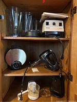 Vita-Mix Super 5000 blender base, multiple blender pitchers with lids, Oster waffle iron, Hamilton Beach hand mixer, Black & Decker handy chopper parts visibles on wooden shelves inside cabinet.