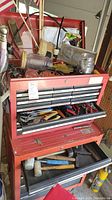 Red metal tool chest with drawers open showing various hand tools inside.
