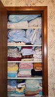 Shelf filled with folded bed sheets, blankets, and pillows in various colors and patterns, showing some signs of wear and stains.