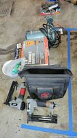Image showing Chicago Electric trimmer router box, two air staplers, black fabric carrying case, and a Fisher Scientific pump with cord and motor in background.