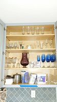 Full view of cabinet shelves showing various glassware including clear stemware glasses, blue stem glasses, a purple pitcher, salt and pepper shakers, drinking glasses, and boxed plate hangers along with napkins.