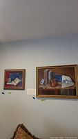 Two framed oil paintings of still life with Jewish ceremonial items hung side-by-side on a wall, showing scale and setting.