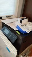 Front angle photo showing Epson WorkForce WF-7840 printer with control panel, numeric keypad, LCD screen, and papers on top including a box of ink cartridges.