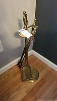 27-inch brass fireplace tool set including shovel, poker, tongs, and brush hanging on a brass stand with circular base, set against a dark wall and hardwood floor.