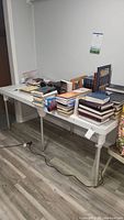 White folding table with metal legs in a room with wood laminate flooring. Various books and items are on the table but contents are not included.