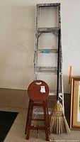 Full view of aluminum ladder, wooden folding stool with round top seat, and broom leaning against wall