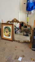 Overall showing large ornate gold toned mirror and leaning signed portrait in red and gold toned frame on floor.