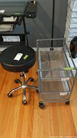 View of the adjustable rolling stool with black padded seat alongside the metal rolling storage cart with mesh drawers