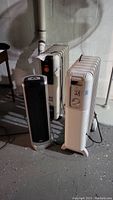 Three space heaters shown together on concrete floor near wall: white Lakewood and DeLonghi oil-filled heaters with black Lasko ceramic tower heater in front.