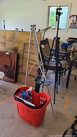Photo showing the aluminum easel, red plastic rolling basket filled with various papers and items, and the Velbon tripod standing behind the basket in a garage setting.