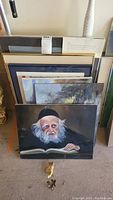 Collection of various framed and unframed art leaning against a wall. Frontmost is a detailed portrait of an elderly man with beard and hat, reading a book.