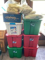 Stack of six plastic storage totes in red, green, and white colors with a cardboard box on top containing packed household items wrapped in plastic bags