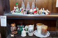 Shelf with an assortment of Christmas figurines including Santa Claus, animated characters, and clear glass Christmas trees on the top shelf, and assorted decorative items including a nesting doll, candle holders, and a milk glass plate on the bottom shelf.