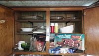 Photo showing wooden cabinet shelves filled with glass casserole dishes, pie plates, bowls, kitchen accessories, and boxed items including SuperThaw wrap and microwave bacon rack.