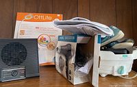 Grouped items photo showing OttLite LED lamp box, Vidal Sassoon hair dryer box with accessories, Pollenex air cleaner, medical shoes, and brace all placed on a wooden surface.