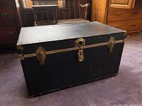 Closed view of the black footlocker trunk with gold hardware, showing scratches and scuffs on exterior.
