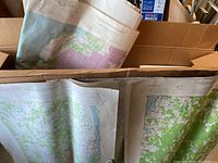 Photo showing several large paper maps with geographic details in green and purple, folded and placed in a cardboard box.