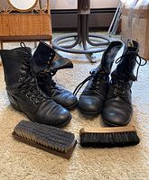 Photo showing the two pairs of black vintage combat boots with laces and two shoe brushes on carpeted floor.