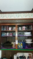 View of cabinet shelf with various mugs, glassware, bowls, plates arranged on shelves with purple liners.