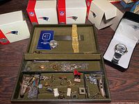 Photo showing four NFL watches in original boxes, one open showing Patriots logo watch. Display tray with various men's jewelry pieces including cuff links, rings, and tie clasps.