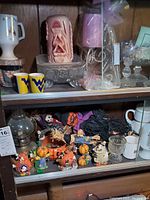 Photo showing two shelves with an assortment of Halloween figurines, candles, an oil lamp, mugs, and glassware including candlesticks and a footed bowl.