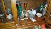 Wooden cabinet shelf with assorted glassware and figurines inside, showing variety of lot items