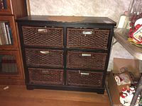 Wooden chest with six rectangular wicker drawers with metal handles, surface showing scratches, positioned near bookshelf and other household items.