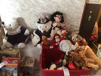 Wide view of assorted stuffed animals and holiday plush on display including larger Luna bears dressed in matching purple velvet outfits and a variety of Christmas-themed plush toys in a red bin and on sofa.