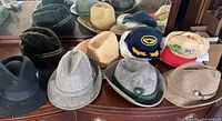Collection of nine vintage men's hats on a wooden surface in front of a mirror. Includes dress hats, ball caps, straw hat, gatsby style hat.
