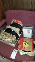 Vinyl 45 RPM records and CDs placed on a red chair, records stacked in boxes and CDs in jewel cases.