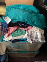 Stack of folded casual women's clothing including teal shorts, pink and light sweaters, blouses, and scarves packed in plastic bins.