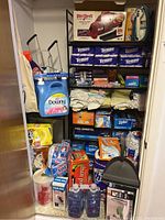 Photo of closet shelf filled with various cleaning supplies and household items including a Dirt Devil vacuum box, multiple tissue boxes, Downy fabric softener bottle, and various bags and cleaning tools.