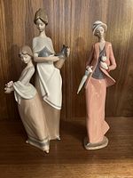 Front view of the two Lladro figurines side by side on a wood shelf with the woman holding pitcher and two women with flowers and tall dress alongside a tall woman in a peach outfit holding an umbrella and cat.