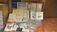 Multiple stamp albums and loose stamps displayed, includes blue 'States Deluxe' album, red binder, white spiral-bound stock book, loose album pages and Ziploc bags with stamps