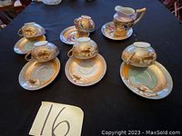 Photo showing four porcelain tea cups upside down on matching saucers, displaying floral and gold leaf designs. Also shows a small pitcher/cream jug with similar-patterned floral decoration.
