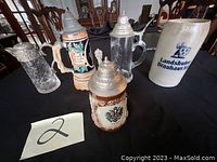 Photo showing five beer steins of varied styles and materials on a table, each with decorative lids.