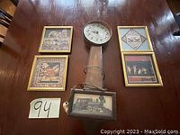 Photo shows four framed pictures and one vintage wall clock placed on a wooden table. The clock has a round face and a long rectangular wooden case with a locomotive picture at the bottom.