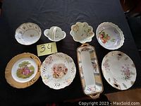 All 8 porcelain plates displayed on dark fabric, showing various shapes, floral and classical designs including pierced edges and a rectangular tray.