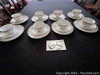Eight white porcelain cups and saucers, each with green garland, pink flowers, and gold trim, arranged on black tablecloth.