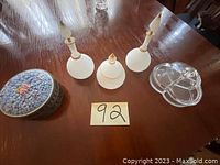 Photo showing all items in the lot arranged on a wooden table: two frosted glass candlesticks, a frosted glass covered jar, a clear sectioned candy dish, and a metal container with a blue lid featuring floral design.