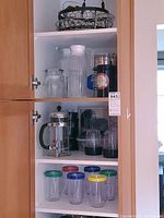 Cupboard shelf with clear plastic canisters with colored lids, Bodum French press coffee maker, glass pitcher, and plastic mugs