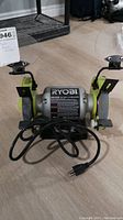 Full view of Ryobi bench grinder showing dual grinding wheels with safety guards and tool rests, power cord in front.